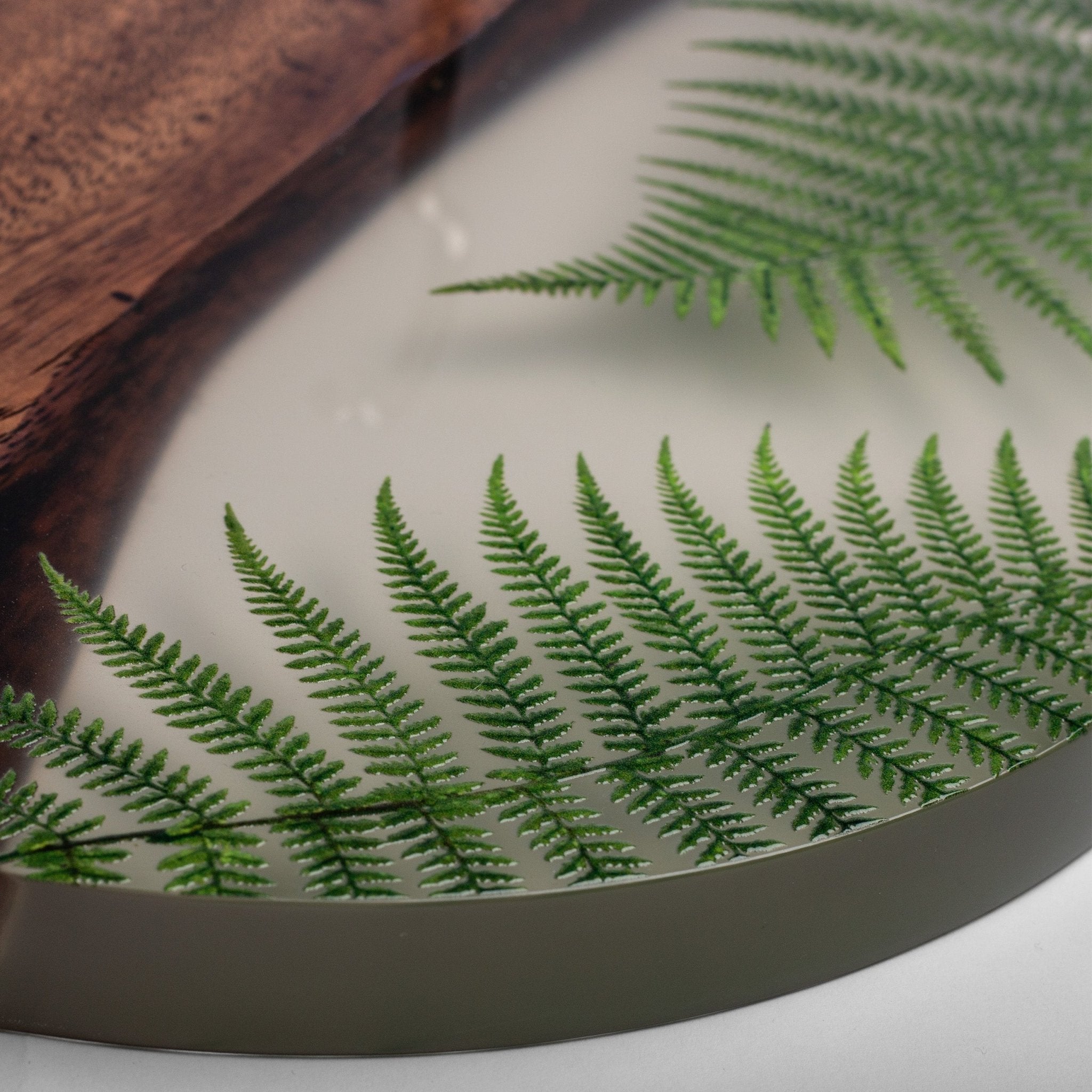 A close-up of a round decorative tray with a dark metallic rim, featuring a light cream center decorated with green fern leaves and a rustic brown wood piece in the upper left corner.
