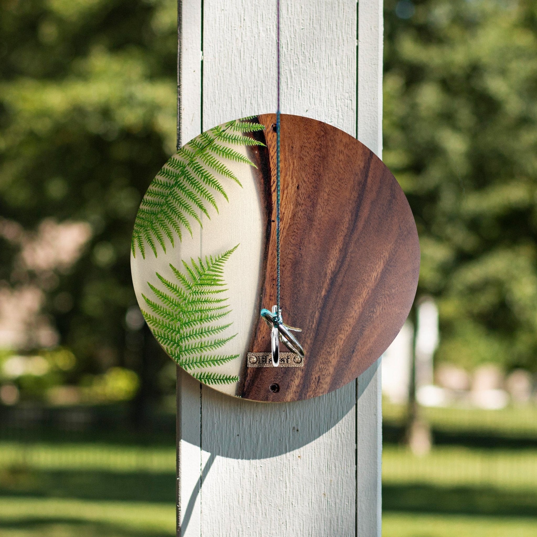A circular wooden wall hanging or decorative plaque with a split design: one half shows a green fern on cream, the other half shows a dark brown wood grain. It hangs from a string in front of a white wooden post with a blurred green outdoor background.