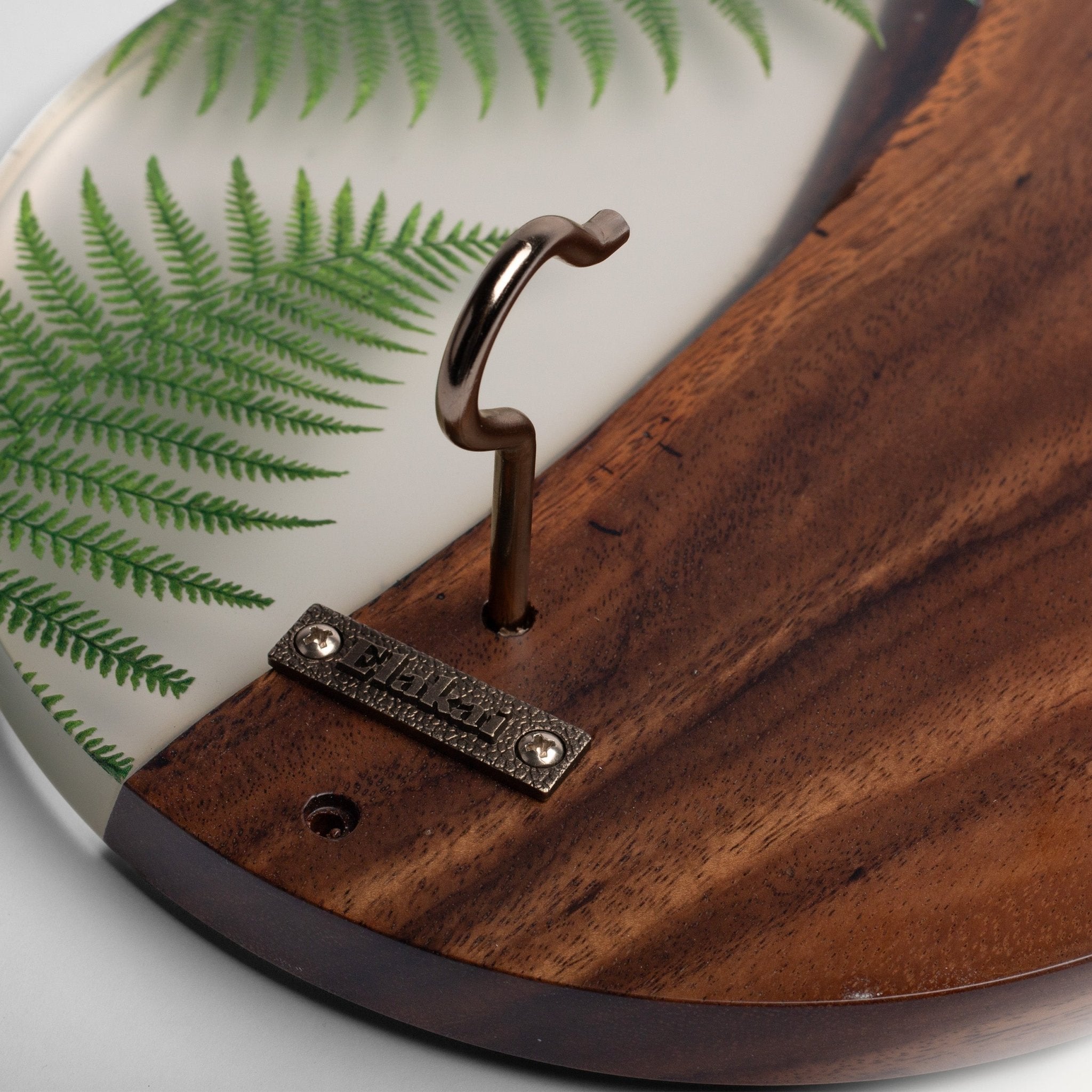 Close-up of a decorative wall hook with a curved dark metal hook mounted on a split design. One half is a rich brown wood grain, and the other half is white with printed green fern leaves.