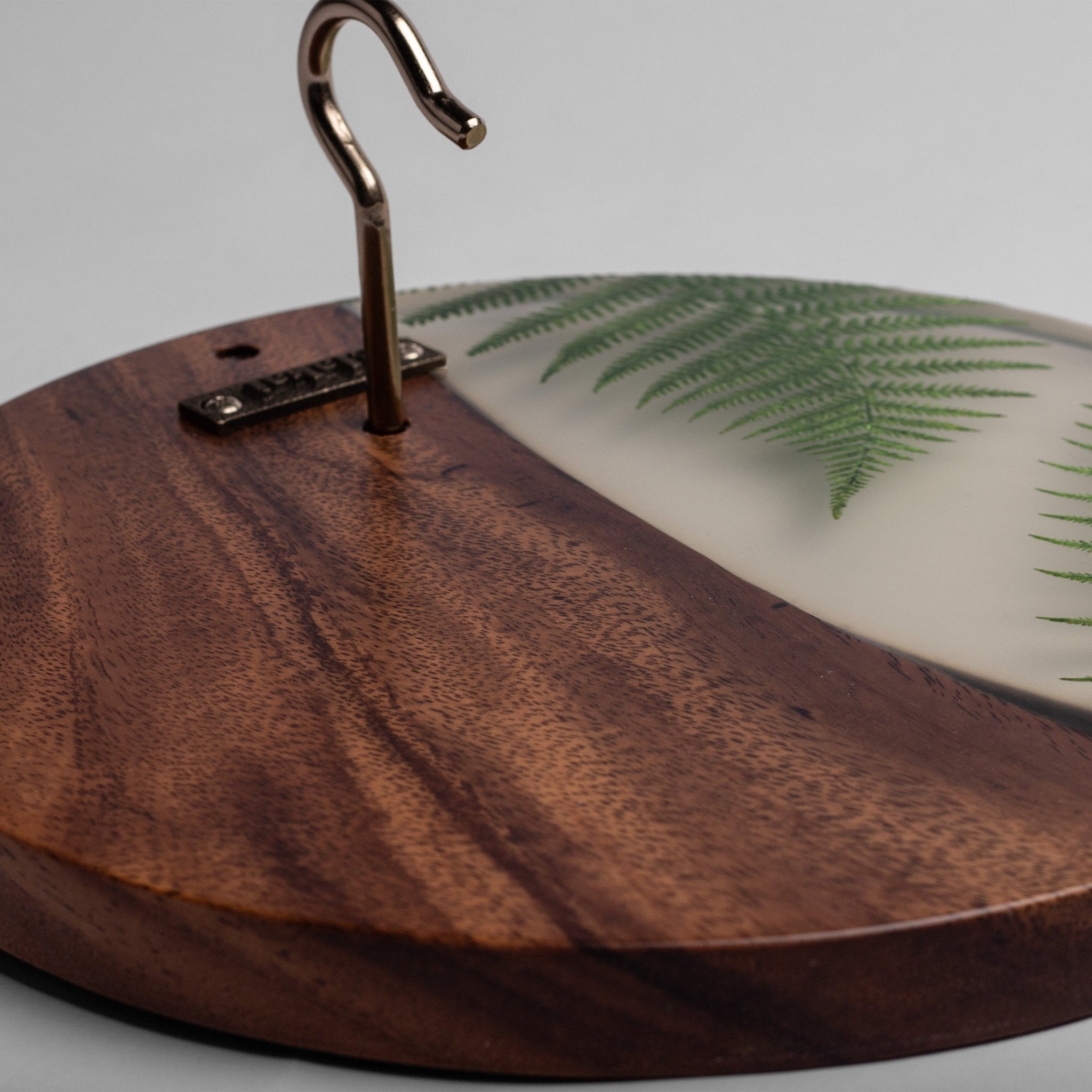 A close-up of a round wooden hanger stand with a dark brown wood-grain finish, a metal hook, and a translucent white inlay featuring a green fern pattern.