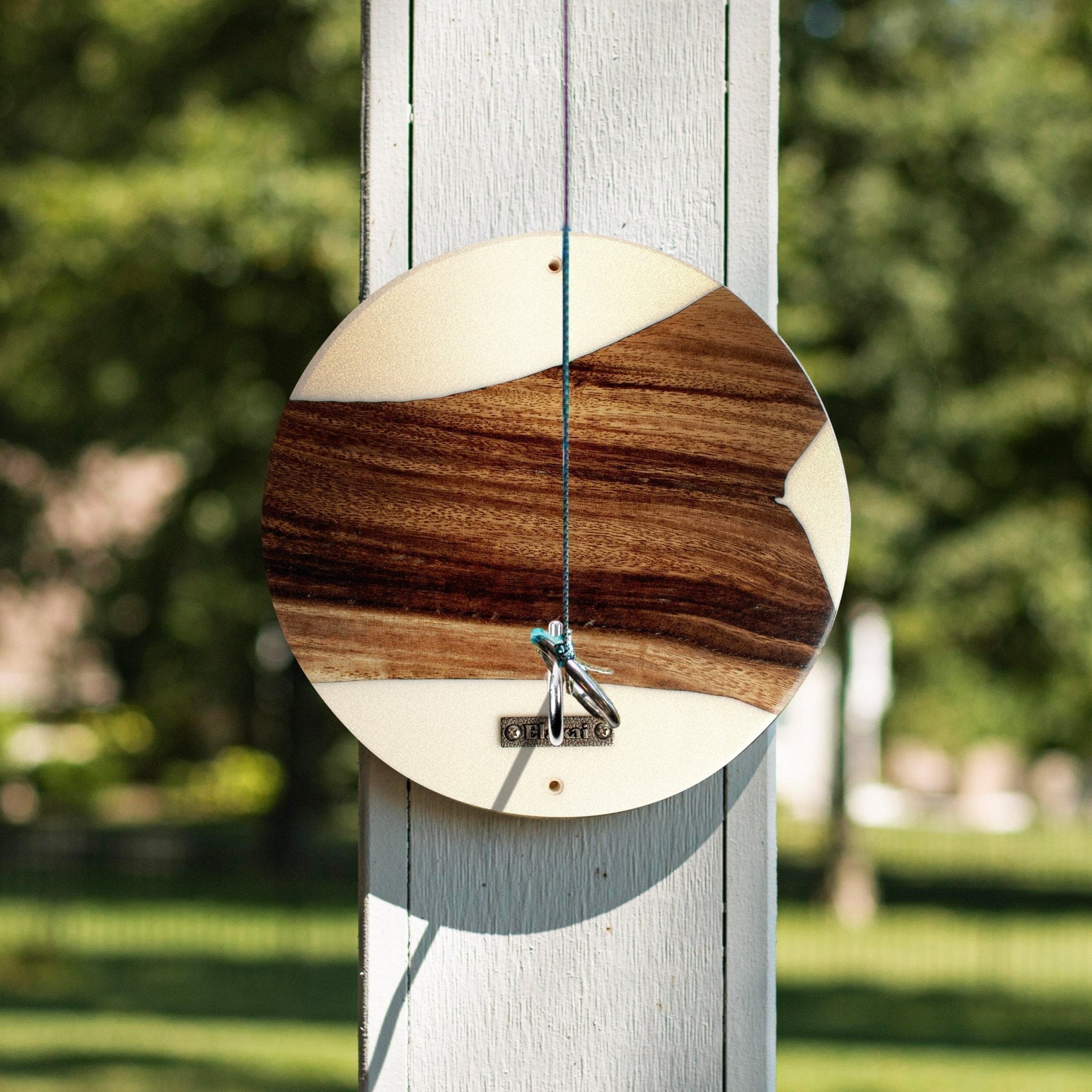 A circular wooden wall clock with a natural wood grain pattern, hanging on a white wooden post in an outdoor garden setting.
