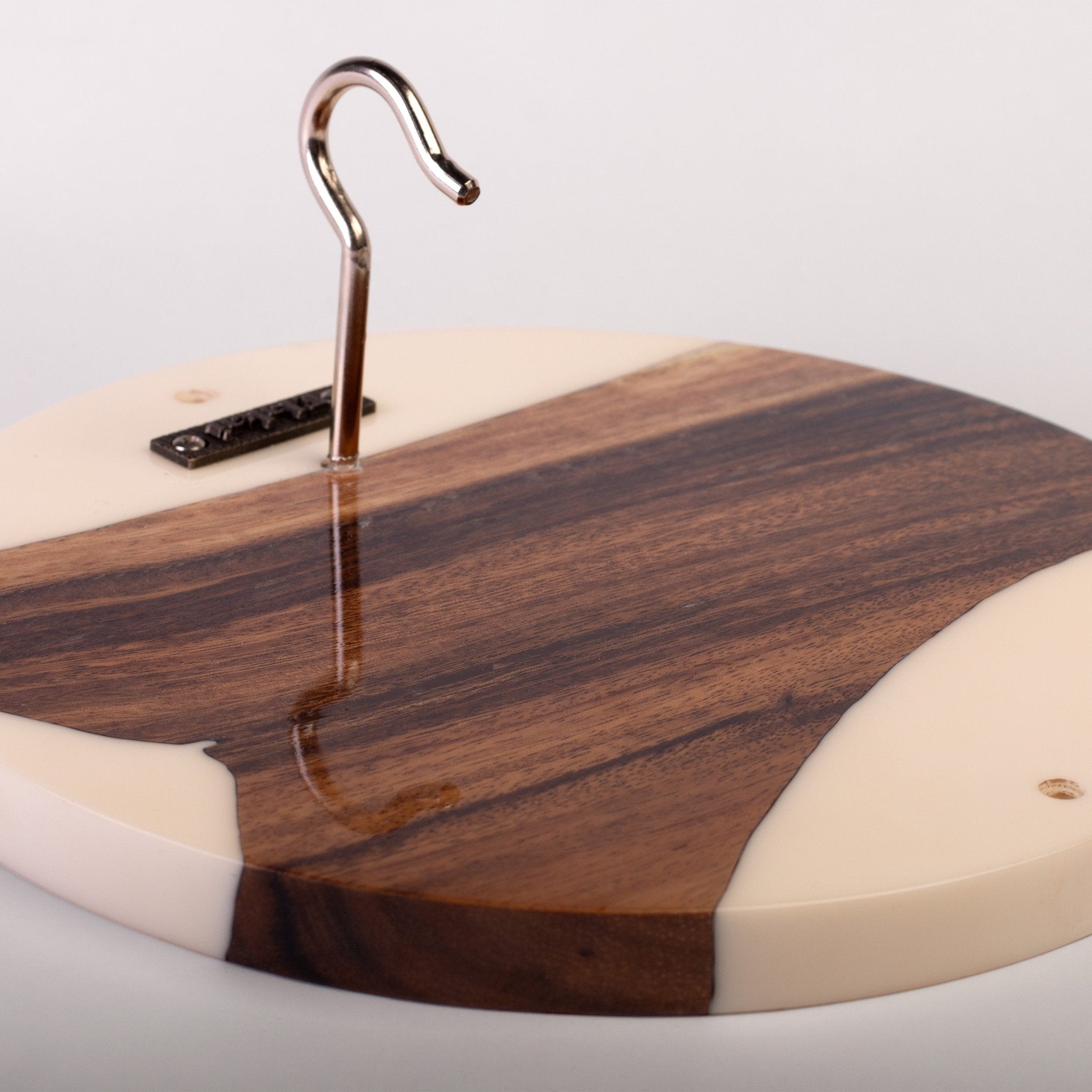 A close-up of a round wooden board with a dark brown wood grain center and cream-colored resin or acrylic sections at the corners. A curved metal hook is attached near the top edge, and a small black rectangular piece is fastened on the left side.