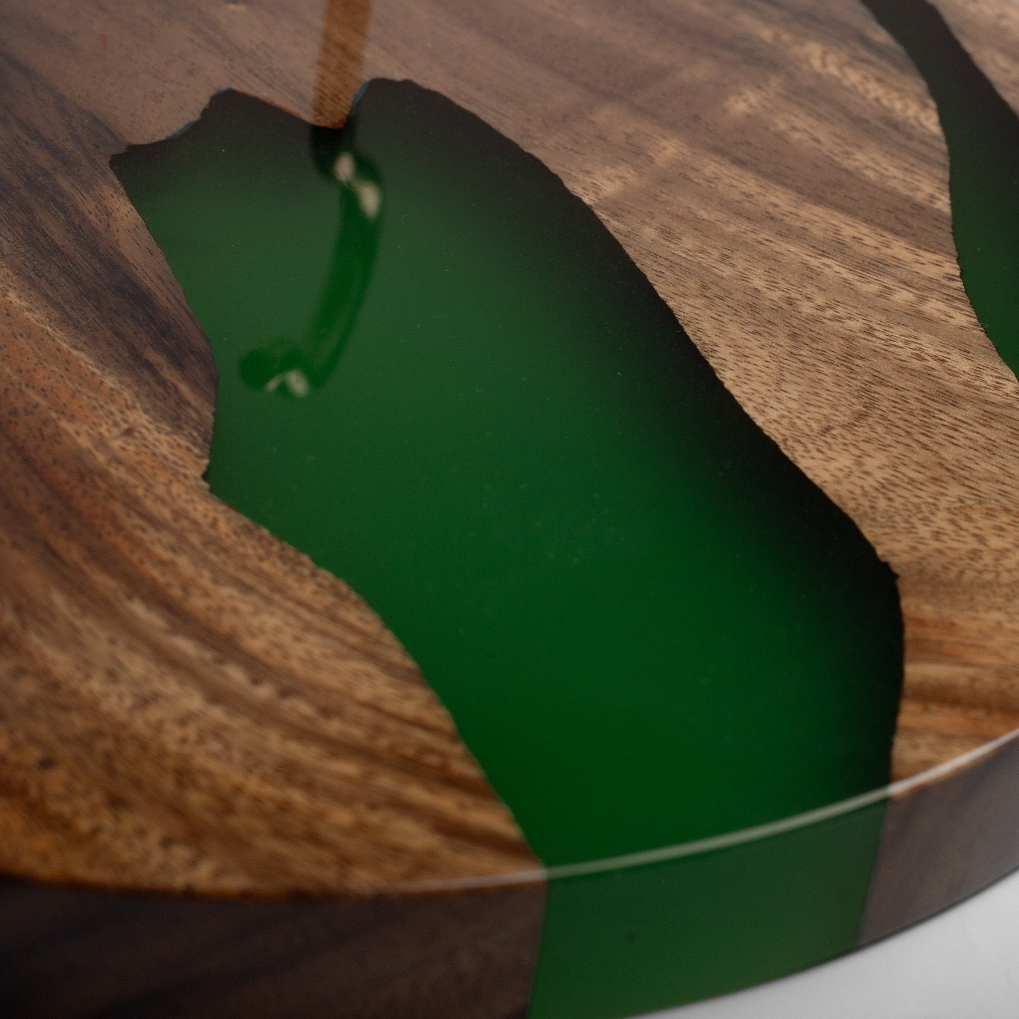 Close-up of a wooden board or tray with a recessed channel filled with glossy dark green resin, showing visible wood grain and a small droplet hanging above the surface.