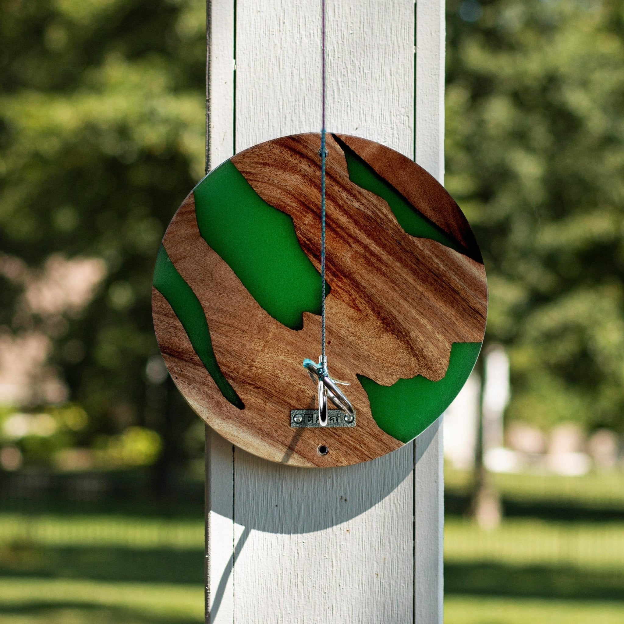 A circular wooden wall clock with green resin inlay and a metal hanging string, mounted on a white wooden post outdoors with a blurred green garden background.