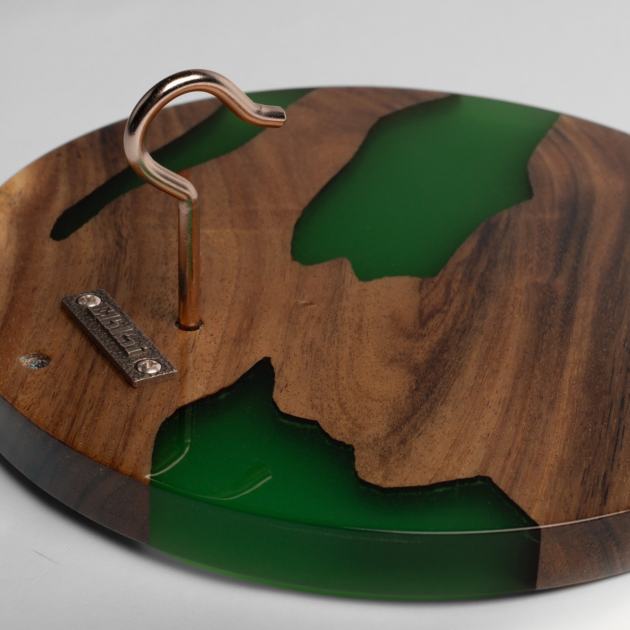 A round wooden plaque or decorative board featuring a cutout map design filled with glossy green resin, with a metal hook hanging from the center and a small engraved brand plate on the side.