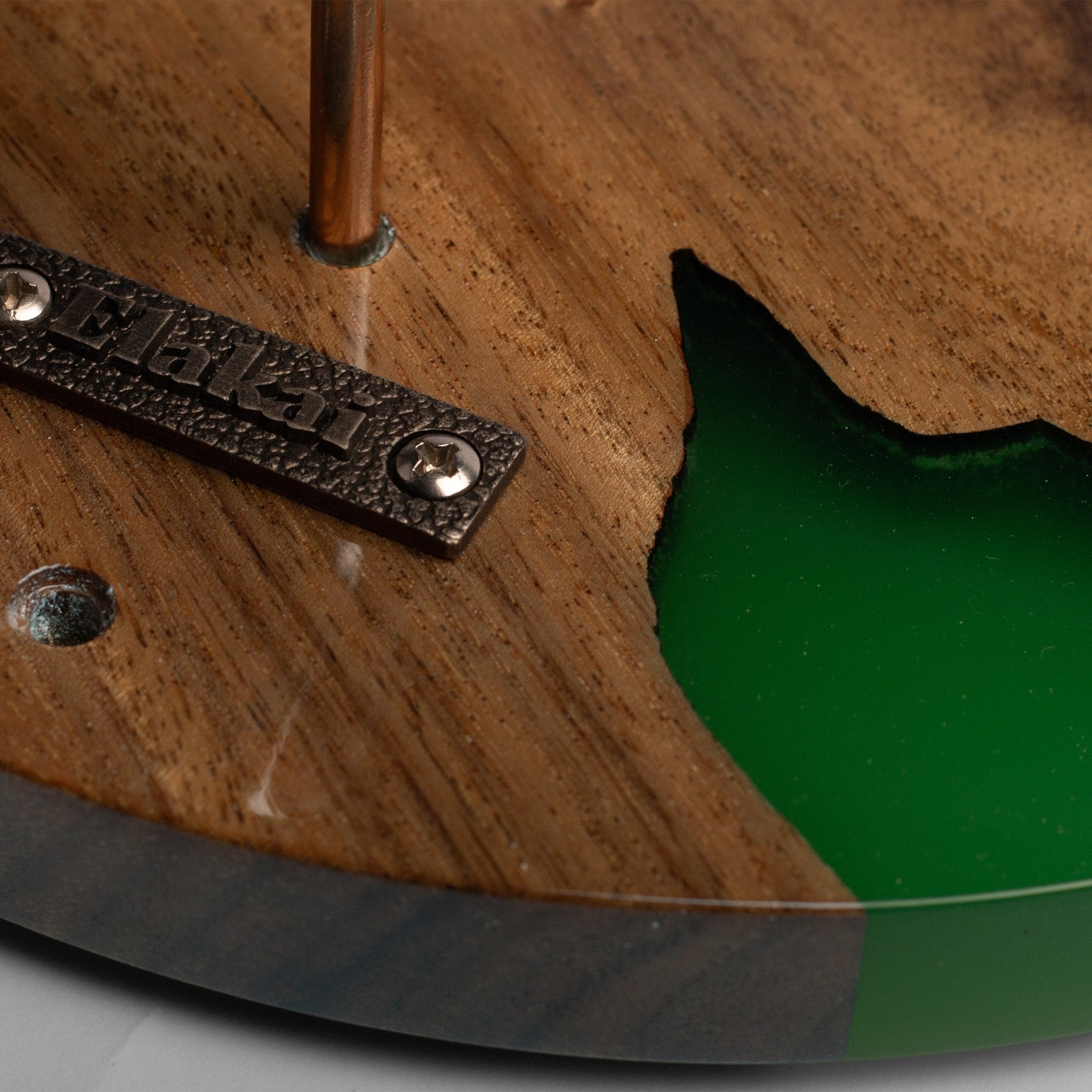 Close-up of a wooden guitar headstock or decorative wooden plaque with a green resin inlay, a dark metal pickguard, and a vertical wood rod.