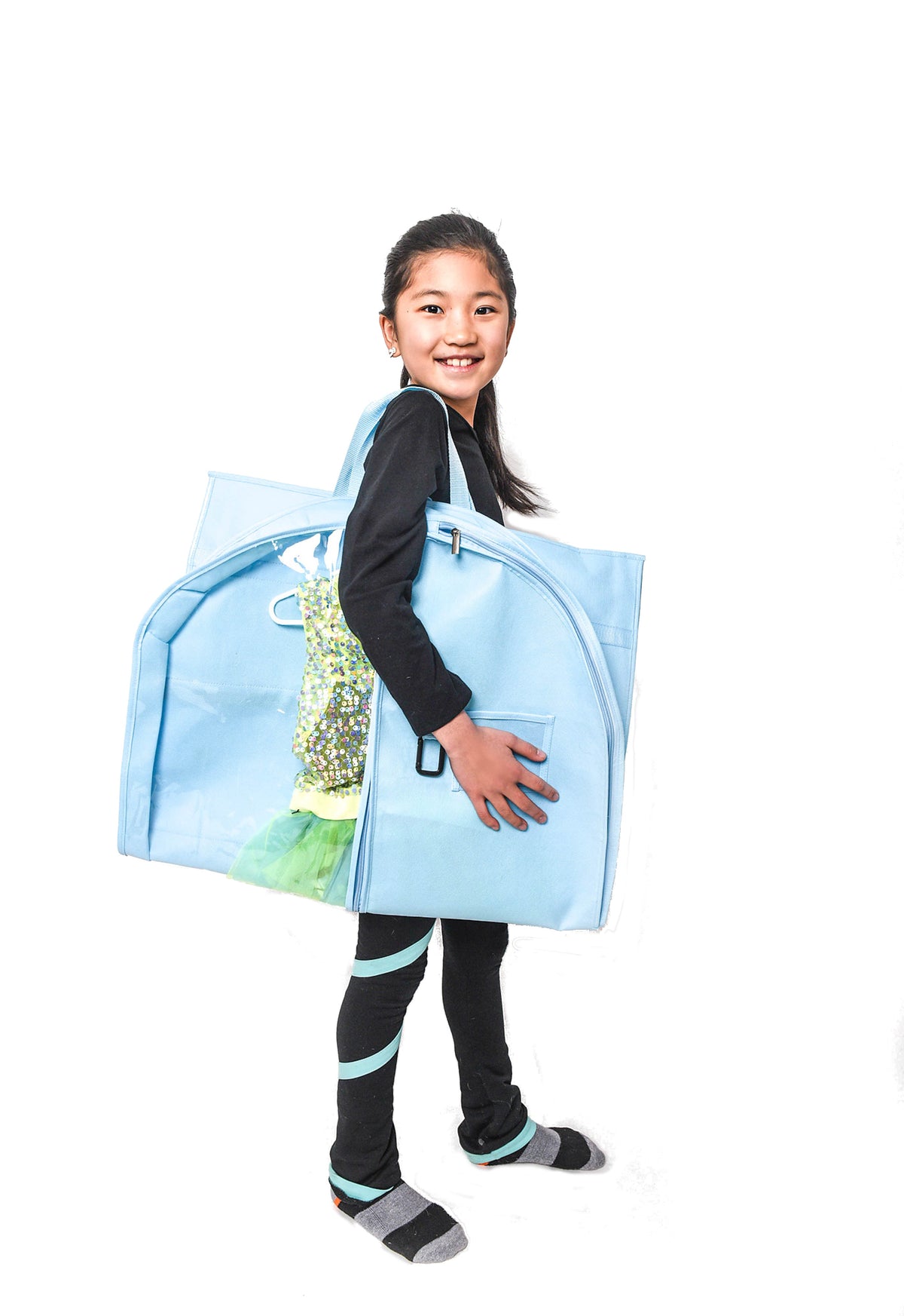 A young girl with dark hair tied back is smiling and looking back over her shoulder while carrying a large, light blue zippered garment bag. The bag has a clear front pocket showing a green and yellow patterned garment inside, and she is wearing black leggings with light blue stripes and gray socks, set against a solid white background.