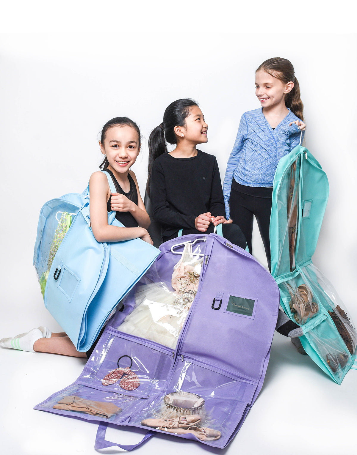 Three children are shown with colorful garment bags in a studio setting. In the foreground, a large lavender garment bag is open, showing clothes inside.