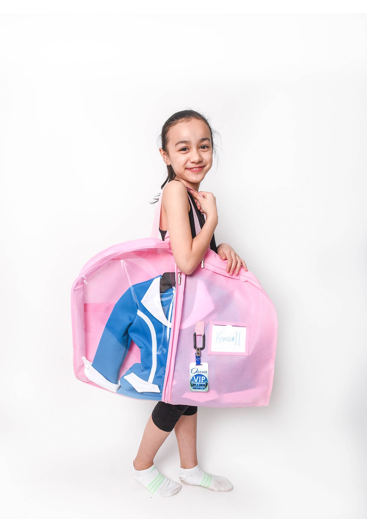 A young girl stands against a white studio background holding a large pink garment bag with a clear front panel. Inside the bag, a blue and white sports jersey is visible.