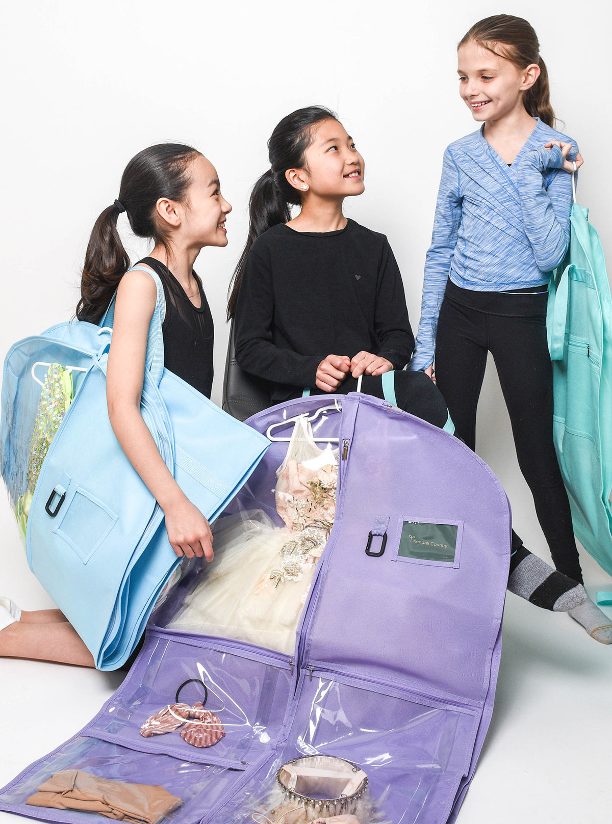 Three girls are shown wearing and carrying light blue dance leotards in a studio setting, with a large lavender garment bag in the foreground holding a white tutu dress.