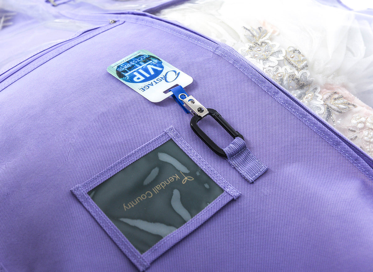 Close-up of a lavender-purple garment bag with a front pocket, a black plastic clip holding a blue and white tag, and a clear window showing a white lace garment inside.