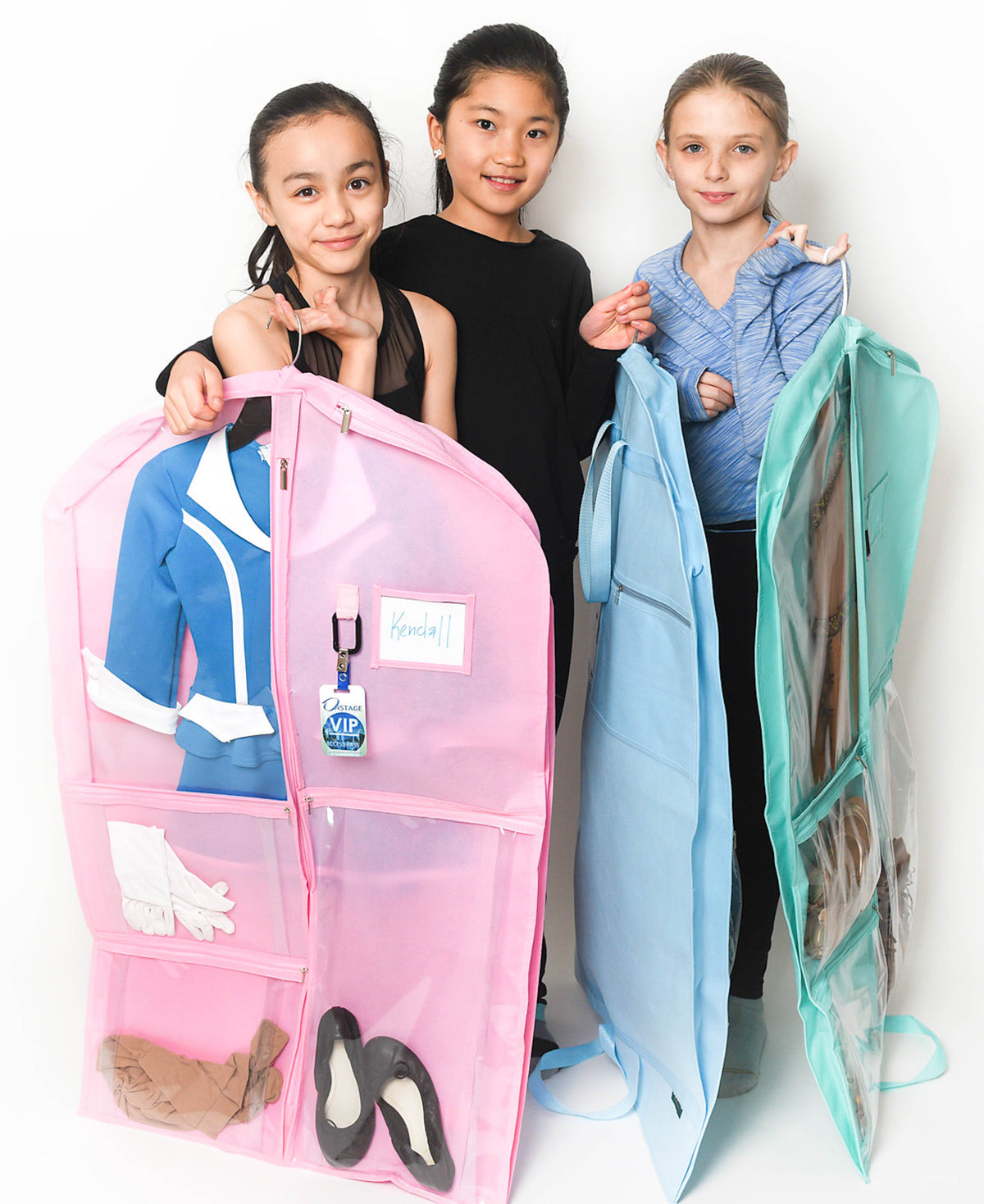 Three girls standing behind two large clear plastic garment storage bags, one pink and one light blue, with clothes and shoes displayed inside on a white studio background.