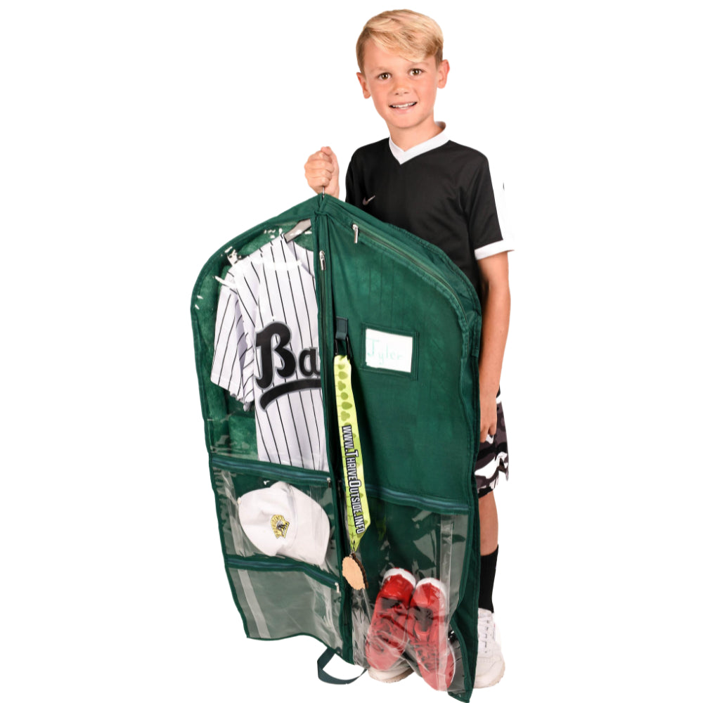 A young boy in a black and white baseball jersey holds up a dark green mesh garment bag filled with sports items, including a baseball jersey, a cap, a bat, and red cleats, against a plain white background.