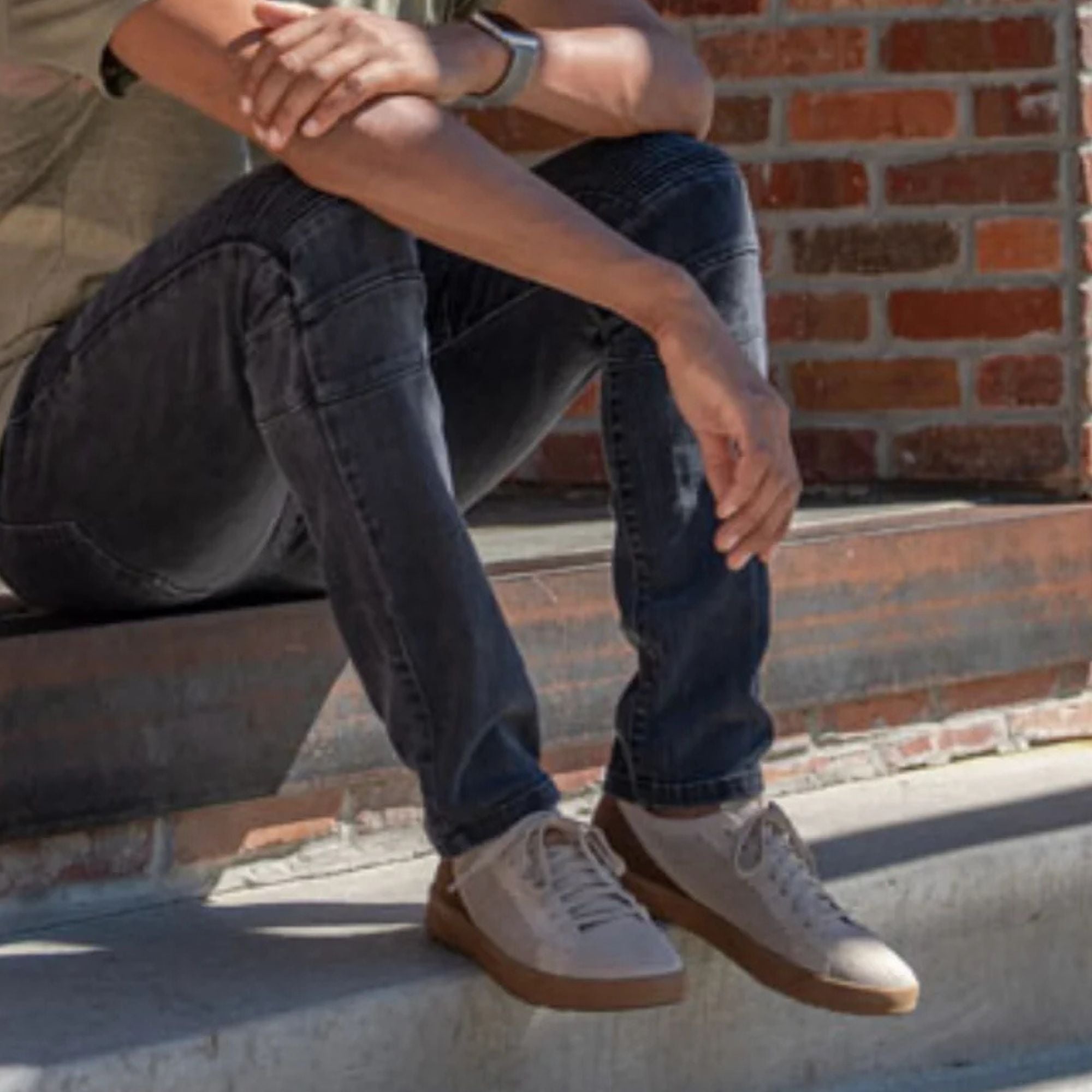 A person sitting on outdoor concrete steps in front of a brick wall, wearing dark gray jeans and beige low-top sneakers with brown soles.