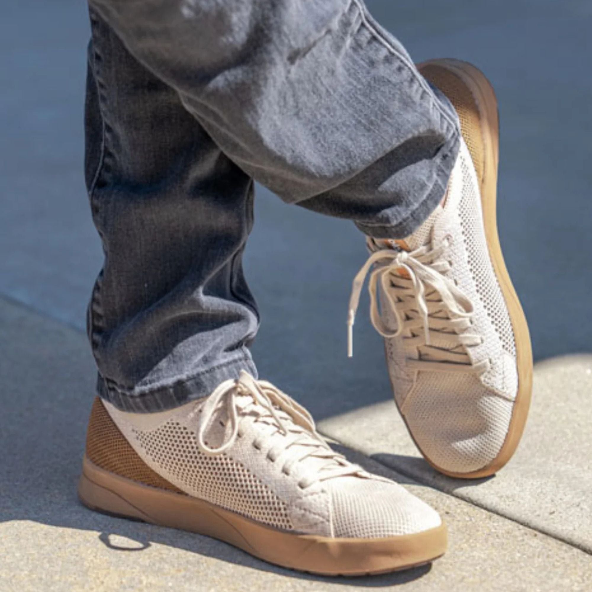 A person wearing off-white mesh sneakers with tan soles and laces, paired with blue jeans, standing on a concrete surface outdoors.