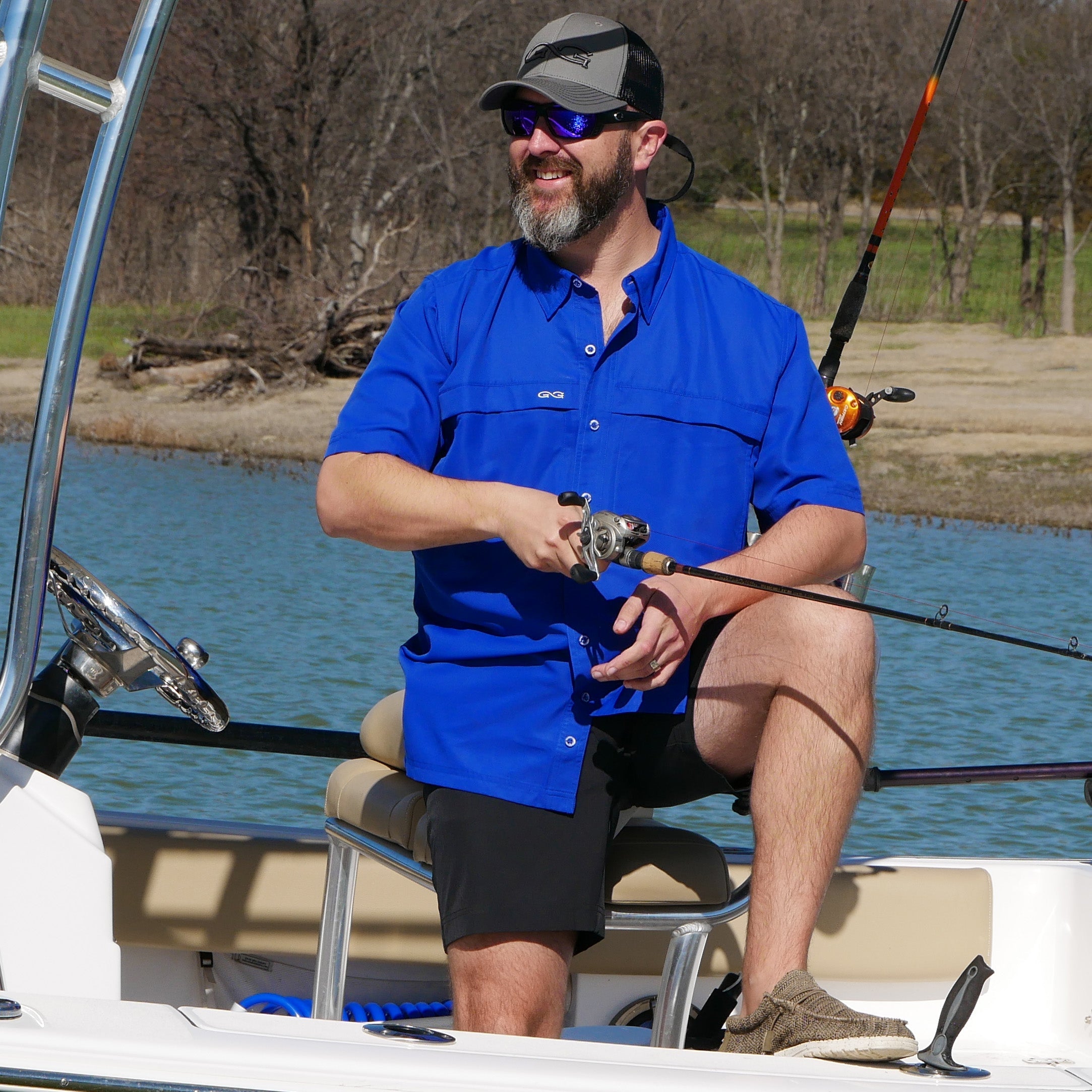 Man in a blue shirt and cap fishing on a boat by a lake.
