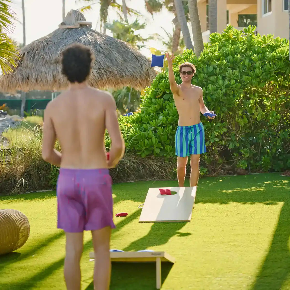 Two men playing a game of cornhole on a grassy area with palm trees in the background.