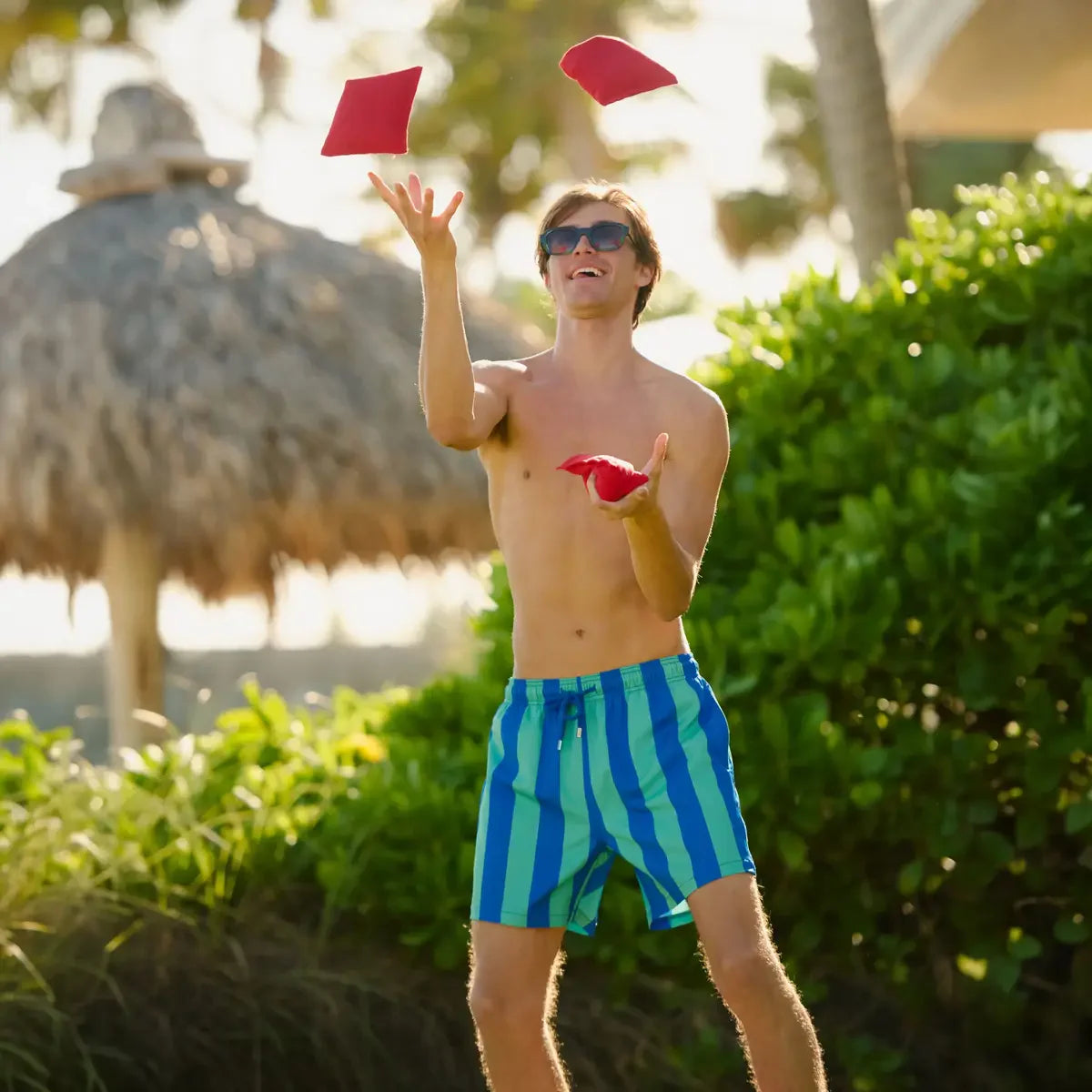 Man juggling red objects in a tropical setting with thatched umbrellas and greenery.