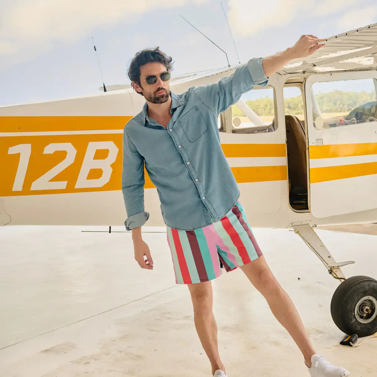 Man in denim shirt and colorful shorts standing next to a small airplane with '12B' on the side.