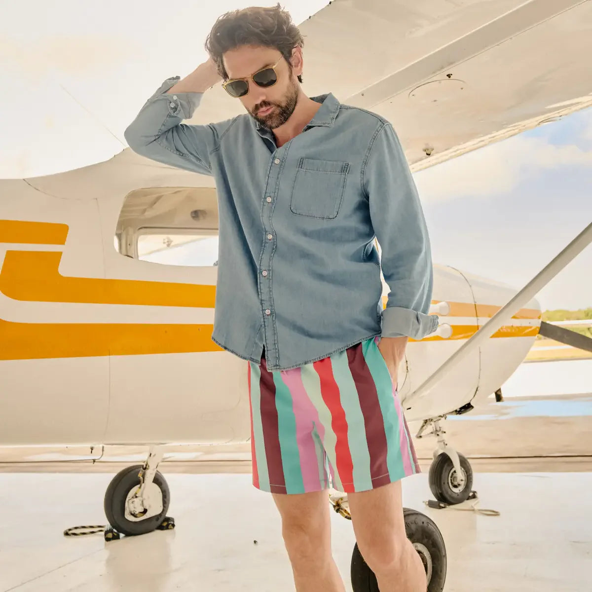 Man wearing a denim shirt and colorful striped shorts standing in front of a small airplane.