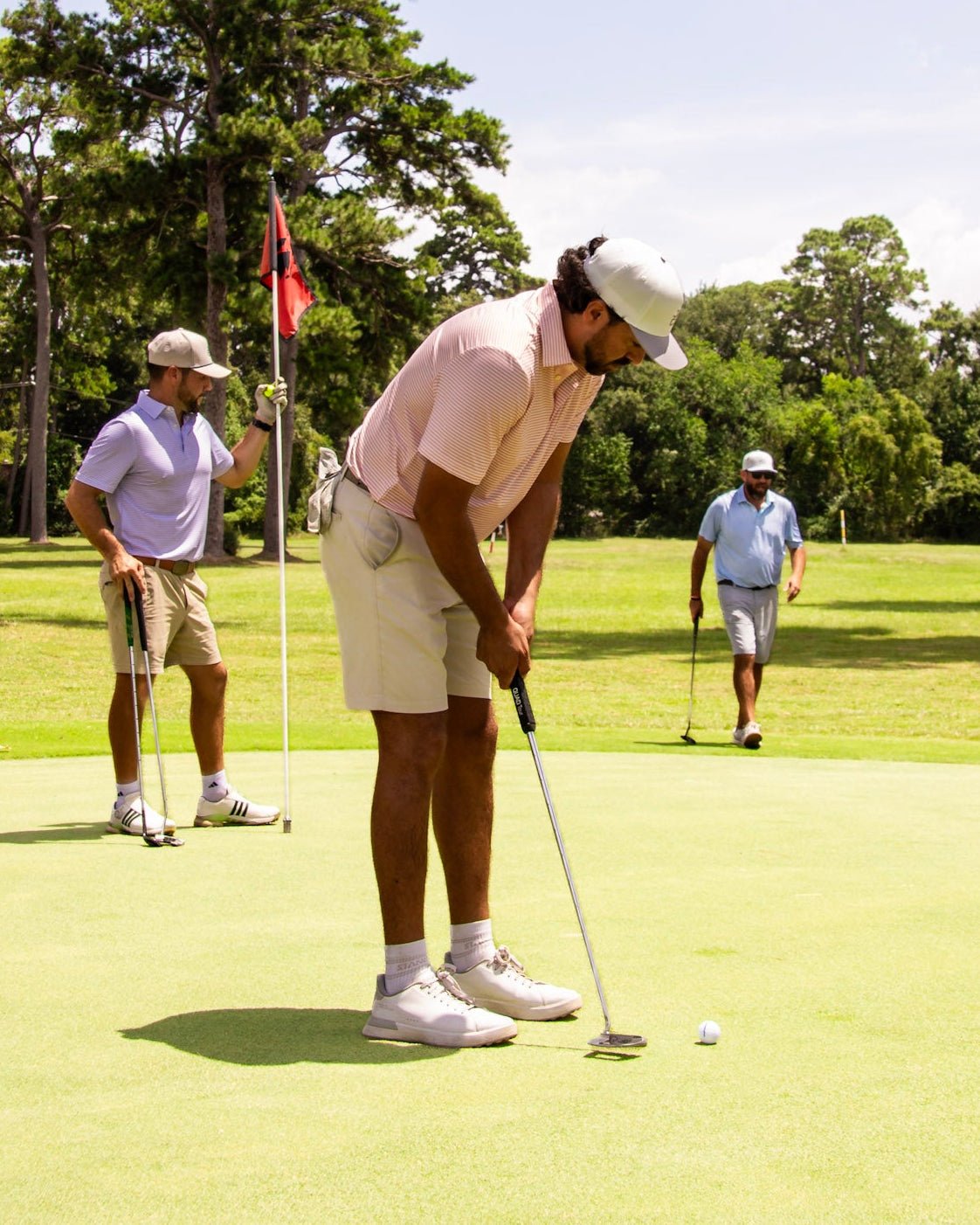 A man in a light pink polo shirt and khaki shorts bends over to putt on a golf green, with another player holding a flag and two other players visible in the background on a sunny day.