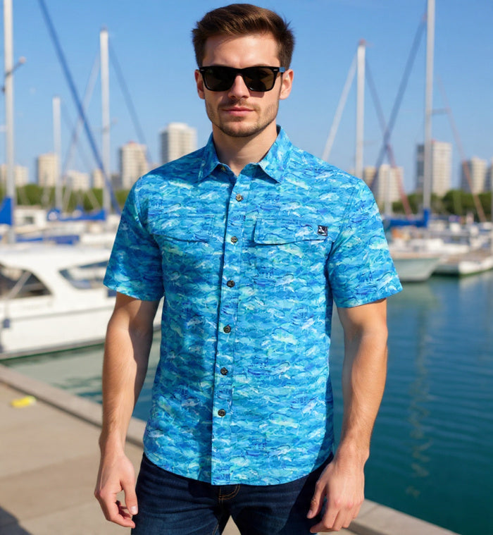 Man wearing a blue patterned shirt standing by a marina with boats and buildings in the background.