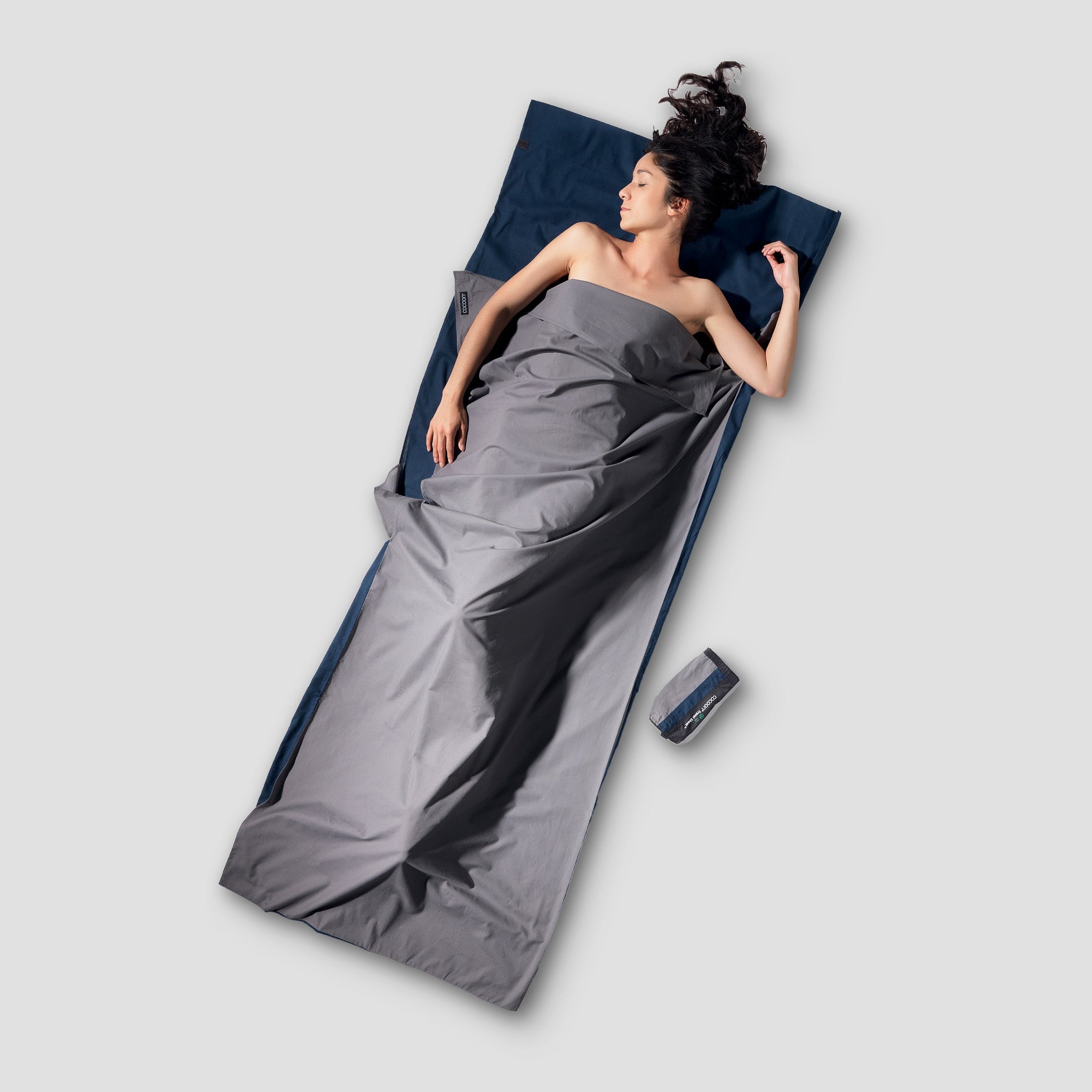 Person lying on a gray sleeping bag with a blue pillow on a light gray background