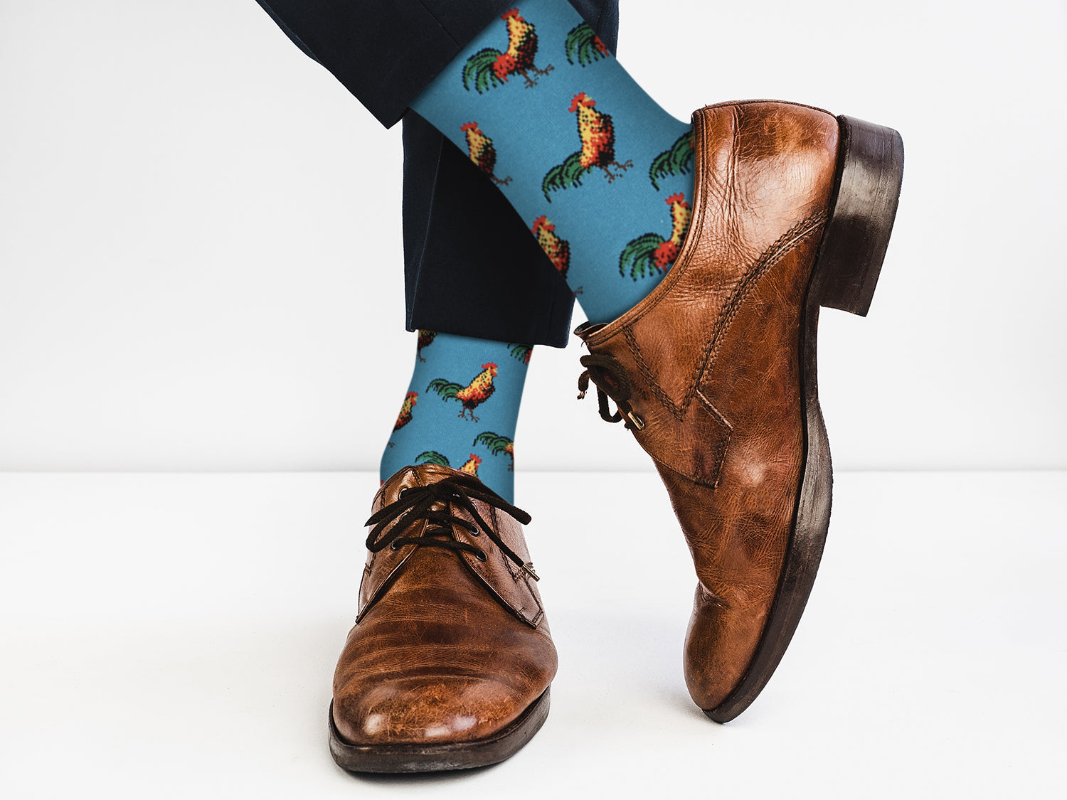 Blue socks with rooster pattern worn with brown leather shoes on a white background