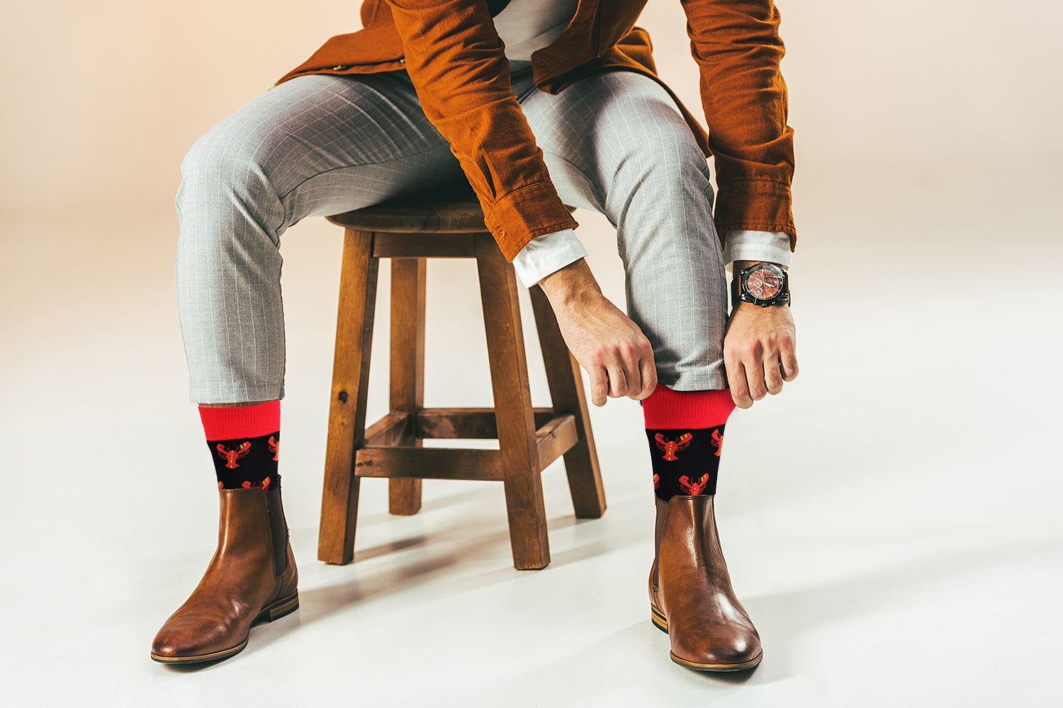 Person sitting on a stool wearing brown boots, light gray pants, and patterned socks with a neutral background.