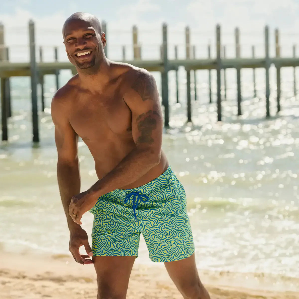 Man wearing green swim shorts with a pattern standing on a beach.