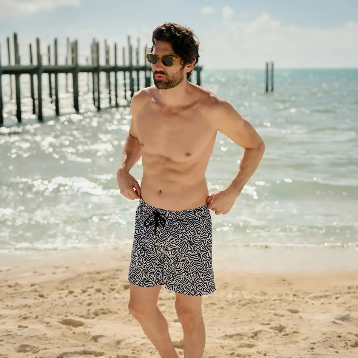 Man wearing patterned swim shorts on a beach with ocean and sky in the background