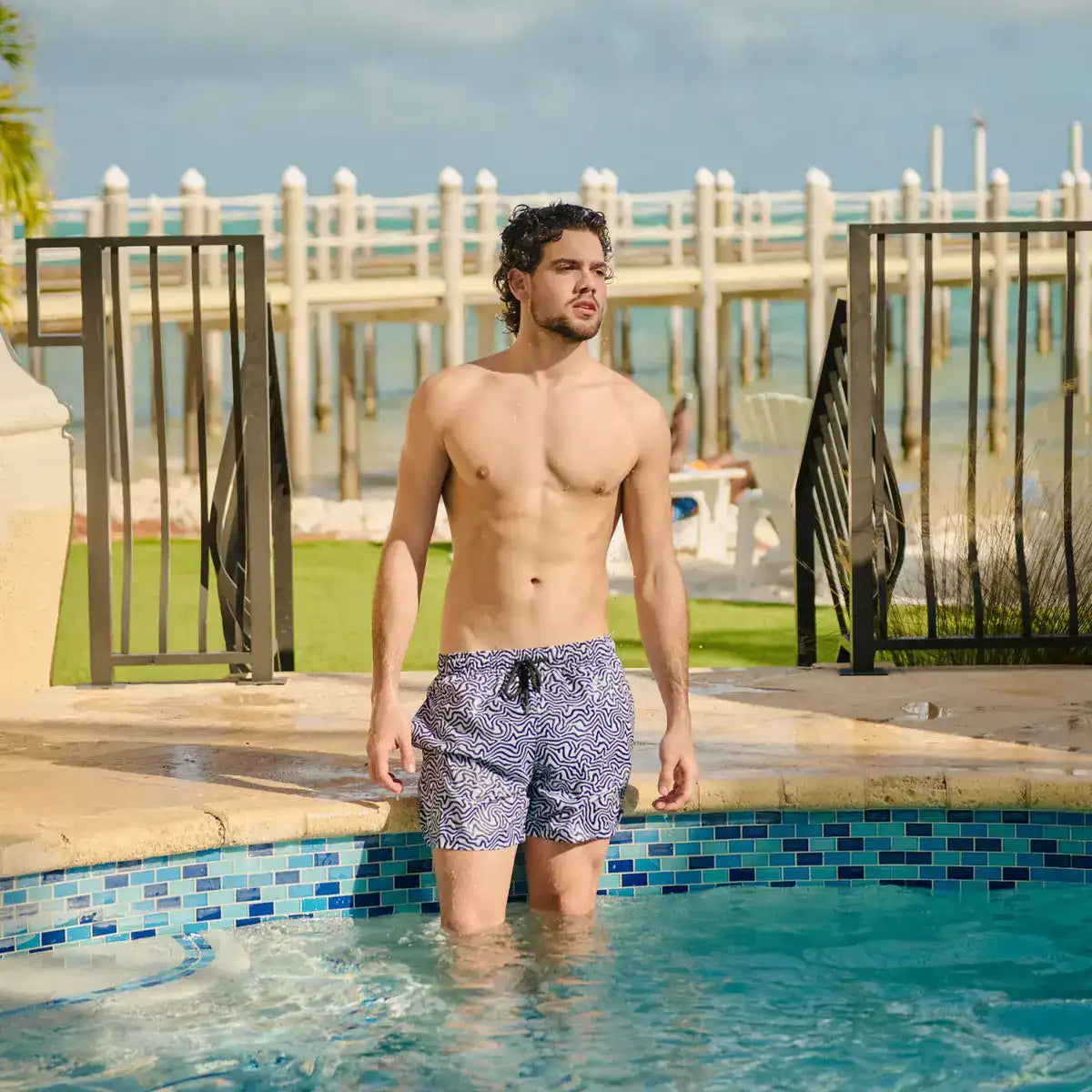 Man in patterned swim shorts standing by a pool with a scenic background