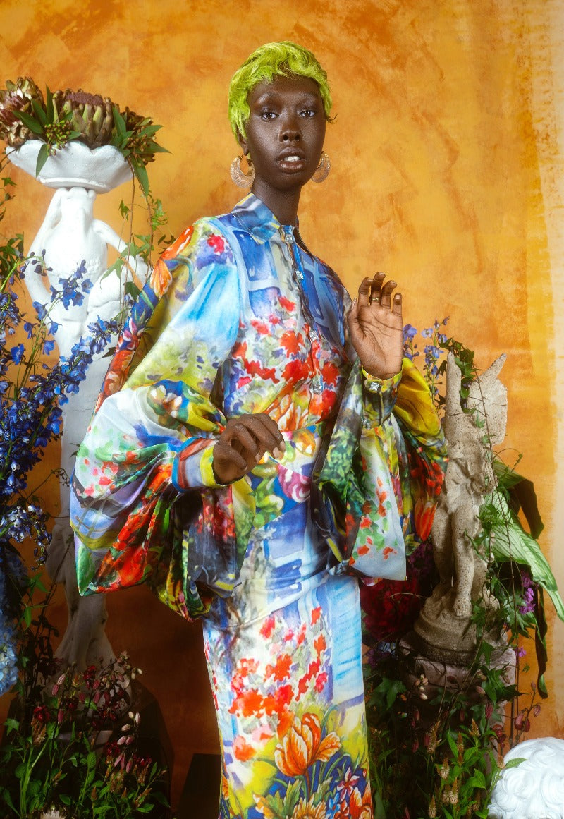 Person wearing a colorful floral outfit standing amidst decorative elements and plants.