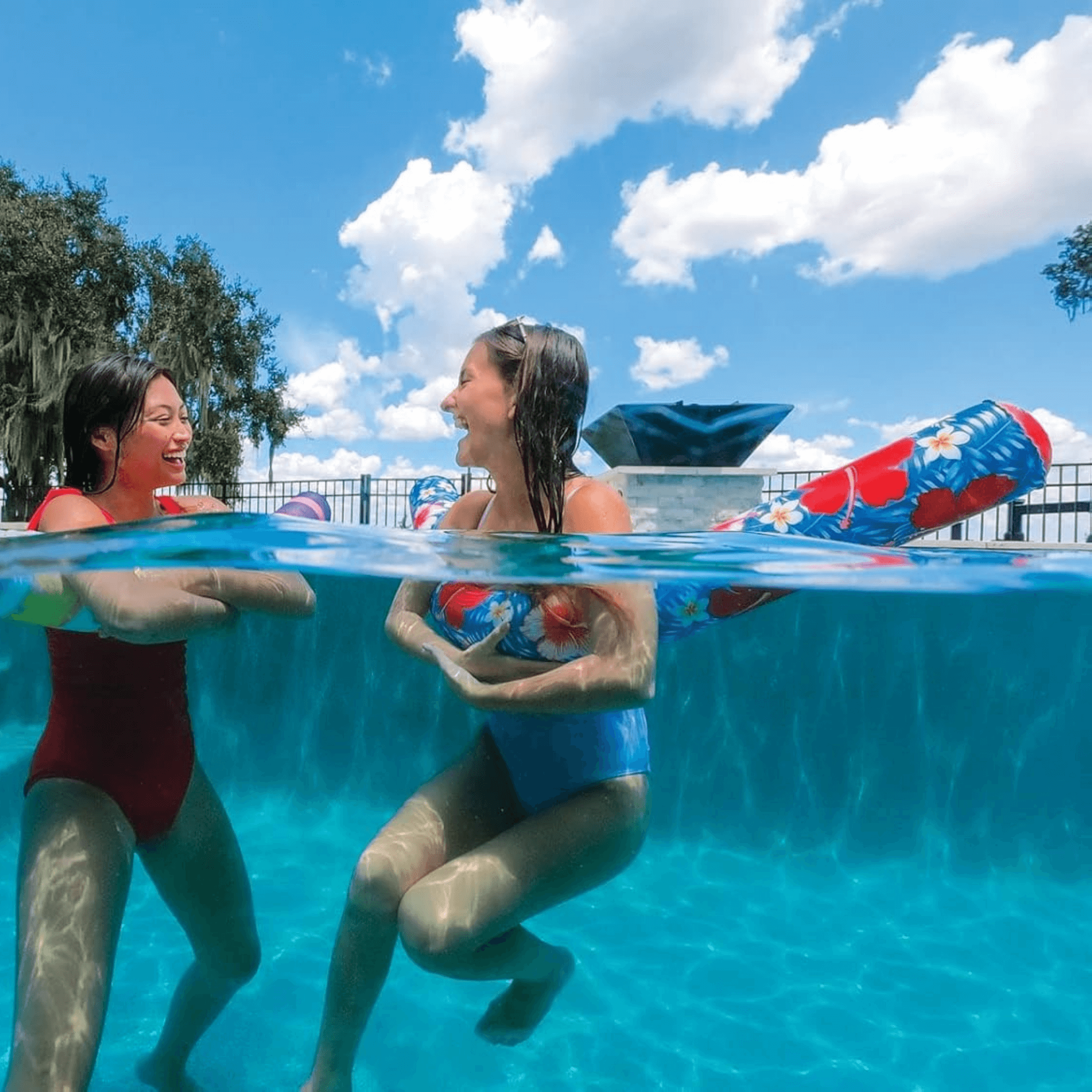 Two women in swimsuits interacting in a pool with a clear blue sky above.