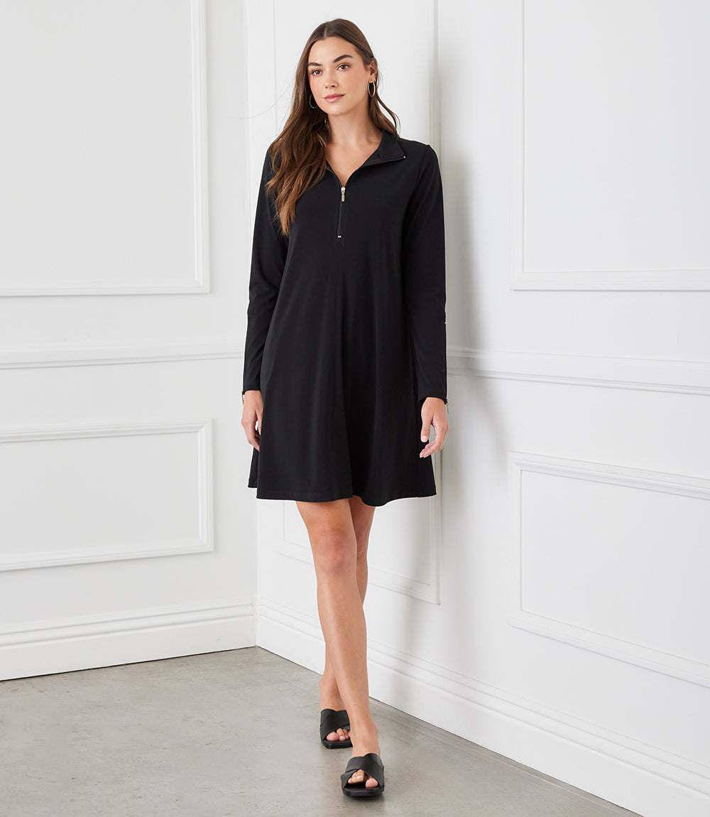 A woman stands against a white paneled wall wearing a black long-sleeve shift dress with a half-zip neckline and black slide sandals.