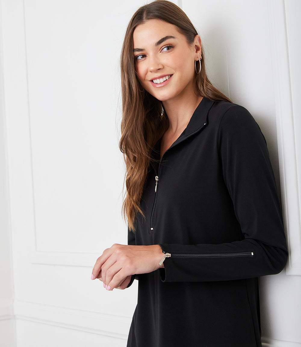A woman with long brown hair wearing a black long-sleeve top with a half-zip collar, leaning against a white paneled wall.
