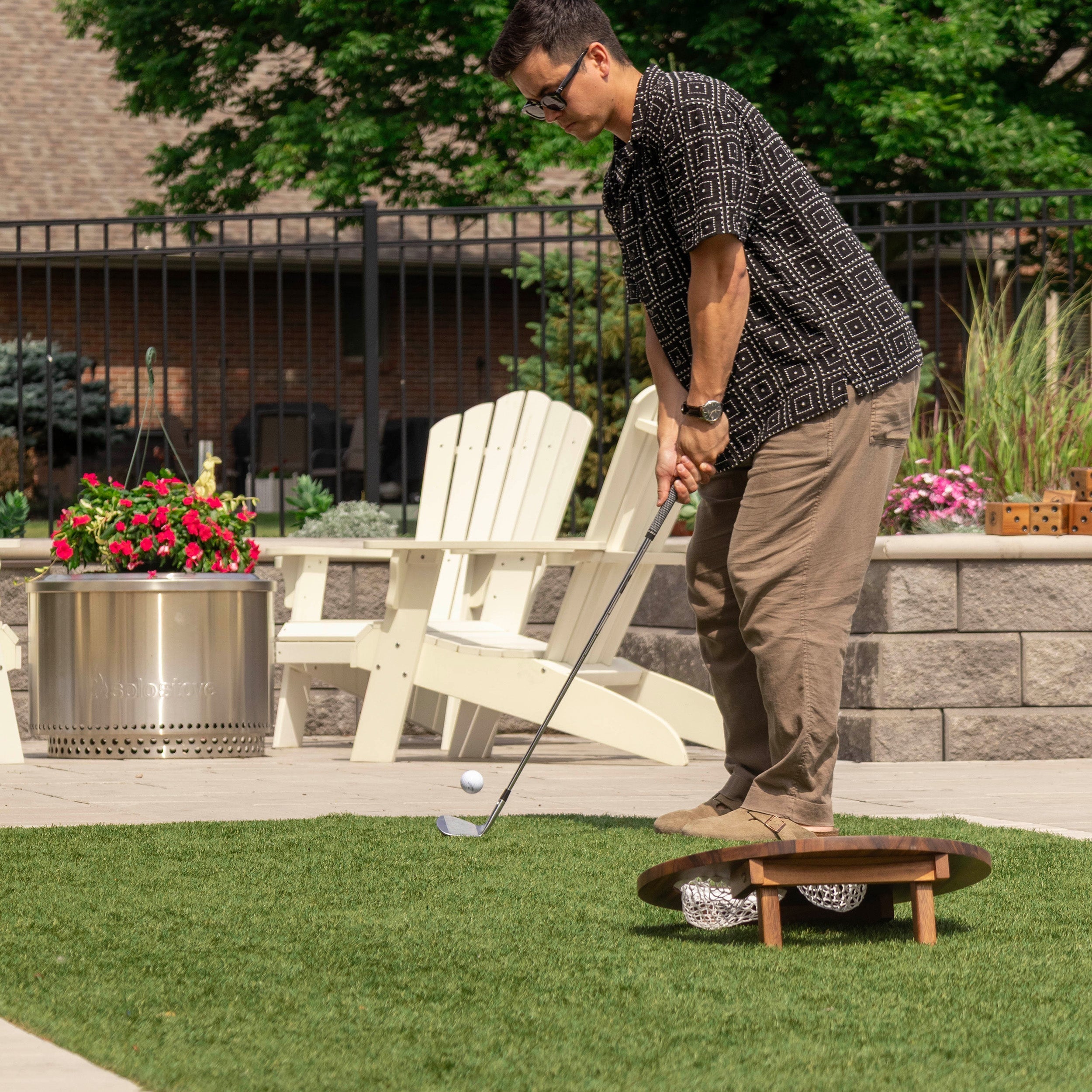 Man playing golf on a putting green with a fire pit and chairs in the background