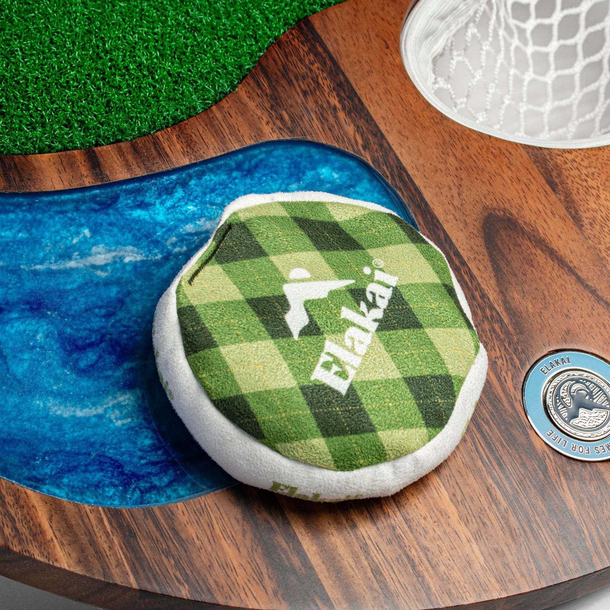 Green checkered Elakat toy on a wooden surface with grass and water background