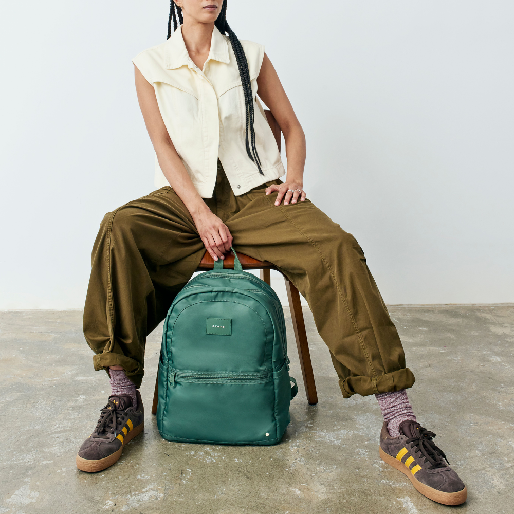 Person sitting on a stool with a green backpack, wearing a cream sleeveless shirt, brown pants, and brown shoes.