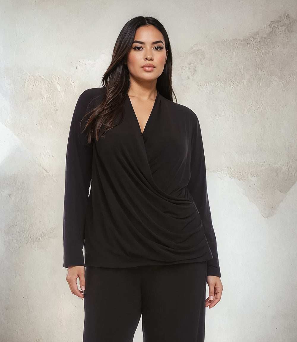 A woman modeling a black long-sleeve wrap-style top with matching black pants, standing against a textured light gray wall.