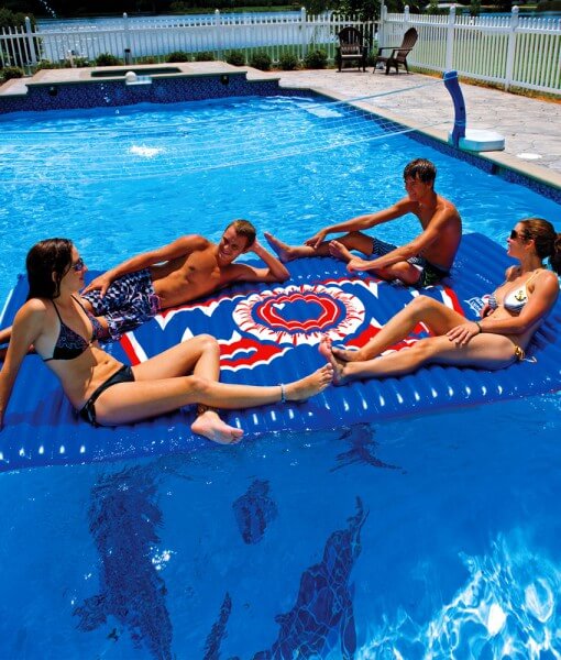 Four people lounging on a large inflatable pool mat with a colorful design in a swimming pool.