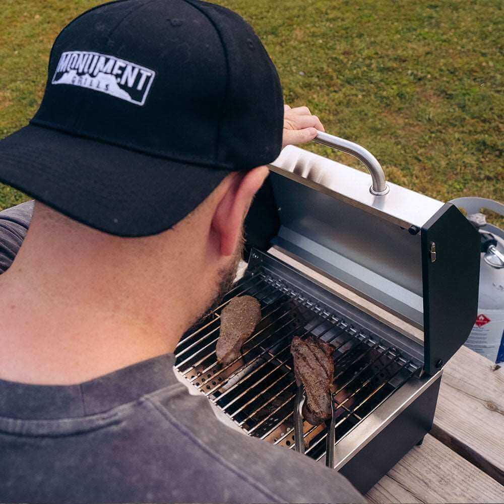 Person grilling on a portable barbecue with Monument Equipment cap