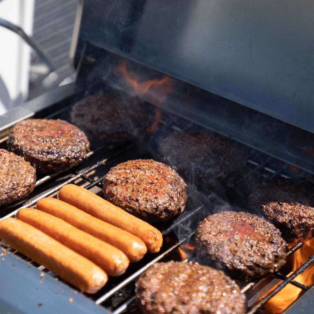Hot dogs and hamburgers cooking on a grill with flames and smoke.