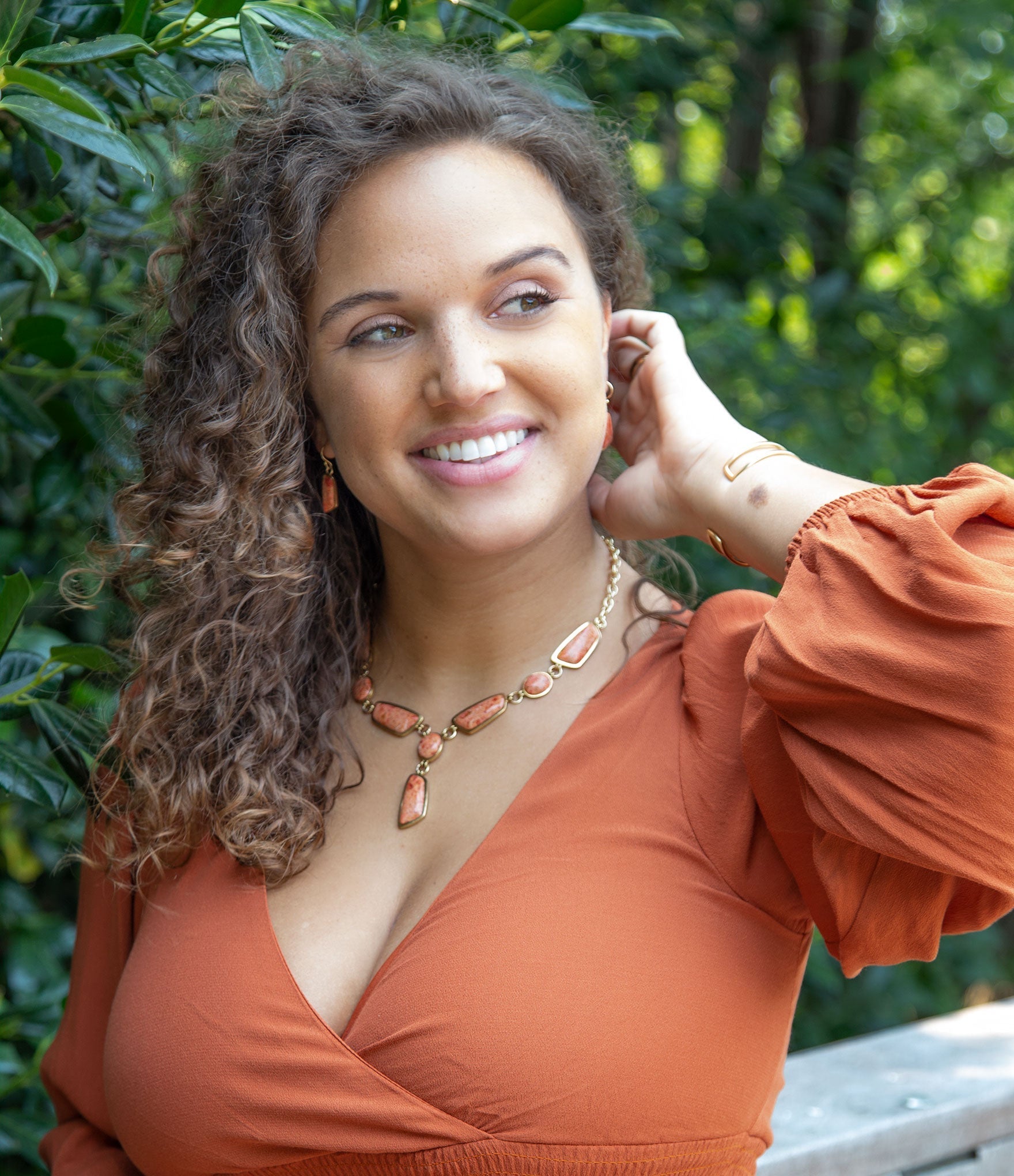 Woman in an orange dress with a necklace, standing outdoors with greenery in the background
