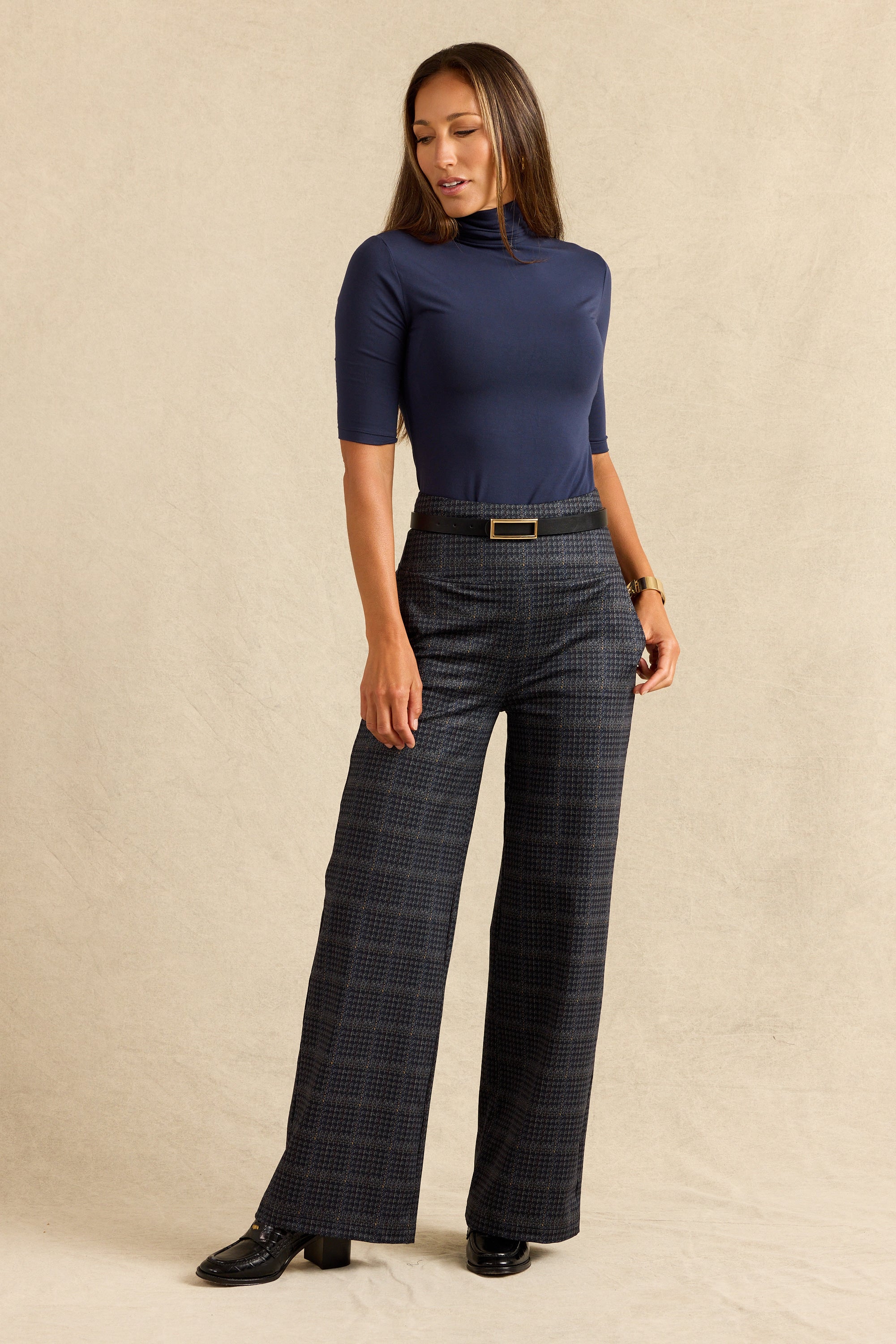 A woman modeling a navy blue short-sleeve mock neck top and dark gray wide-leg plaid trousers with a gold-tone belt, standing against a textured beige studio backdrop.