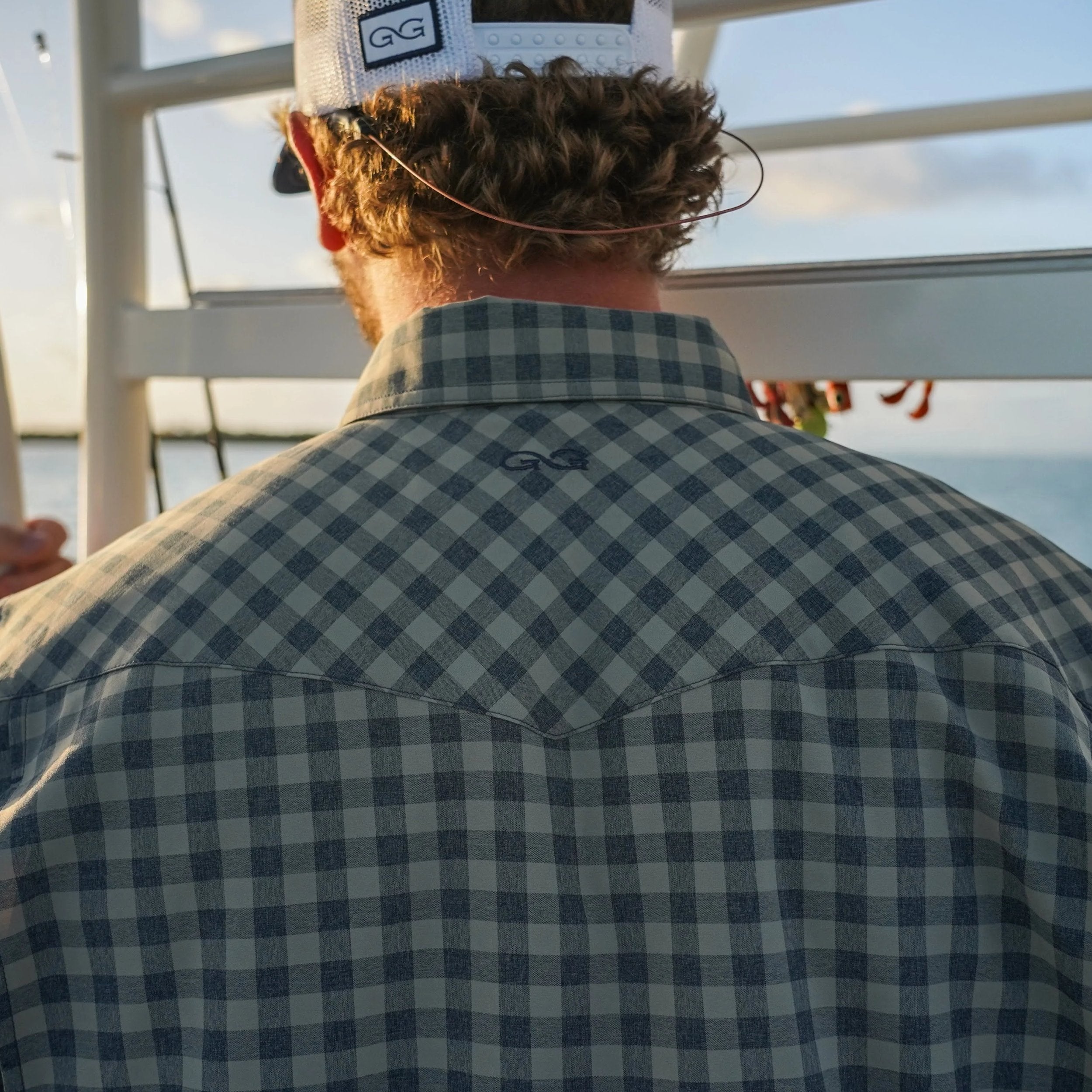 Man wearing a checkered shirt and cap with 'GG' logo, standing on a boat.