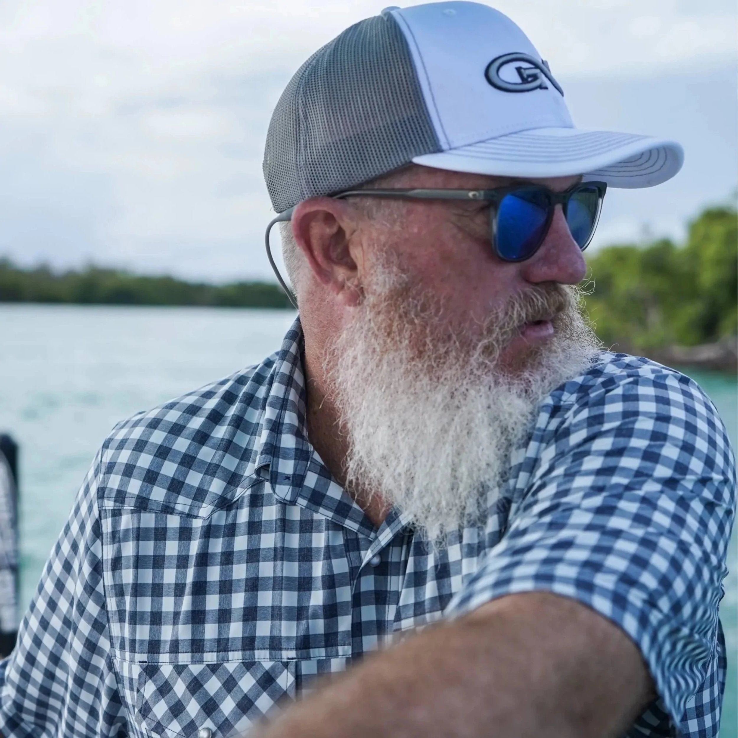 Man wearing a checkered shirt, cap, and sunglasses by a body of water.