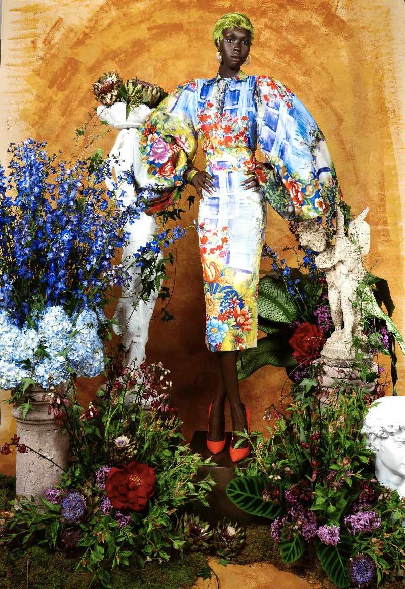 Person in a colorful outfit surrounded by flowers and a statue on a warm-toned background