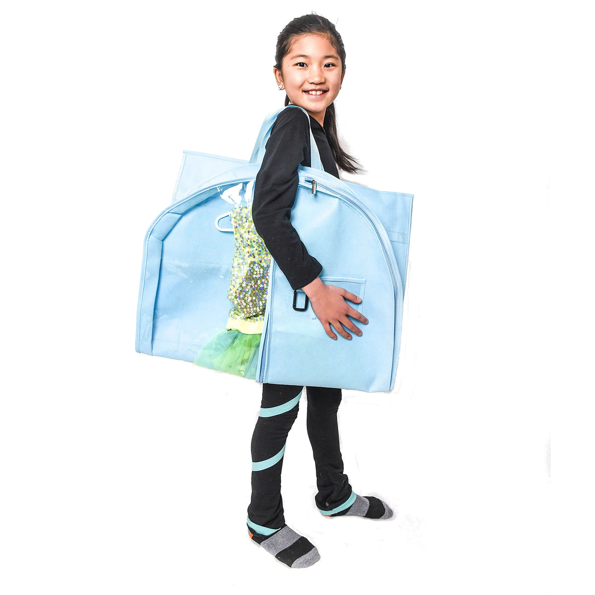A young girl with dark hair tied back wears a black long-sleeve shirt, black leggings with mint blue stripes, and gray socks. She is smiling and holding a large, light blue expandable garment bag filled with a green and yellow patterned dress, set against a solid white background.
