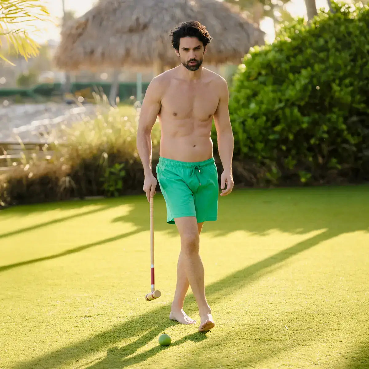 Man in green swim shorts holding a croquet mallet on a grassy lawn with a thatched umbrella in the background.